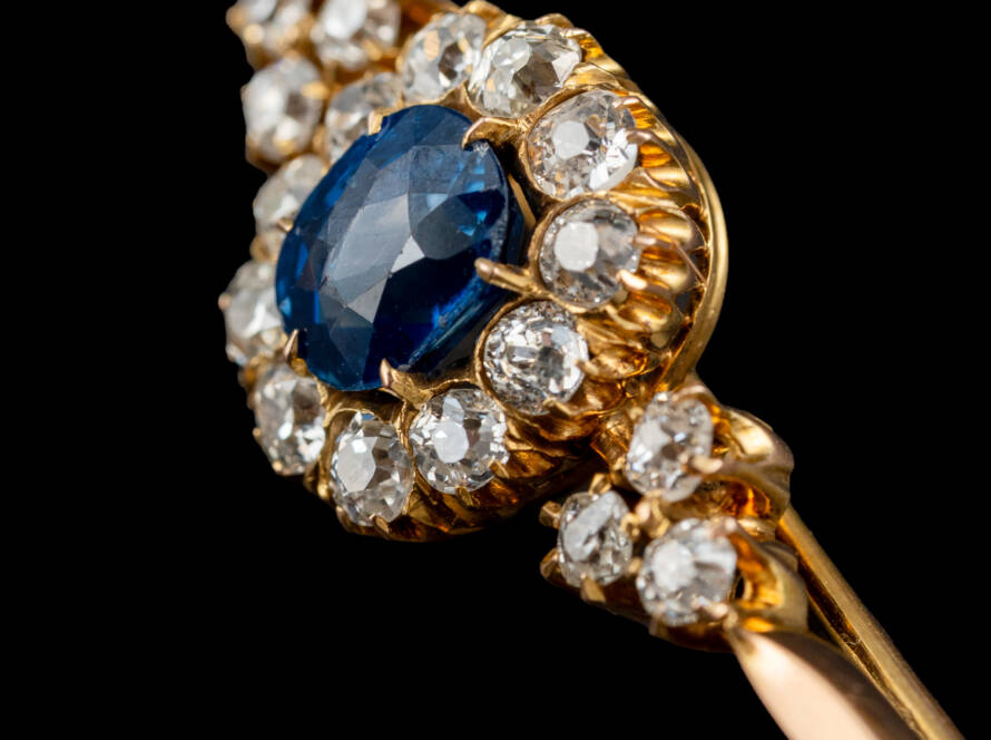 Dettaglio di una spilla vintage in oro con pietra blu centrale e pietre bianche incastonate, fotografia di gioielli su fondo nero
