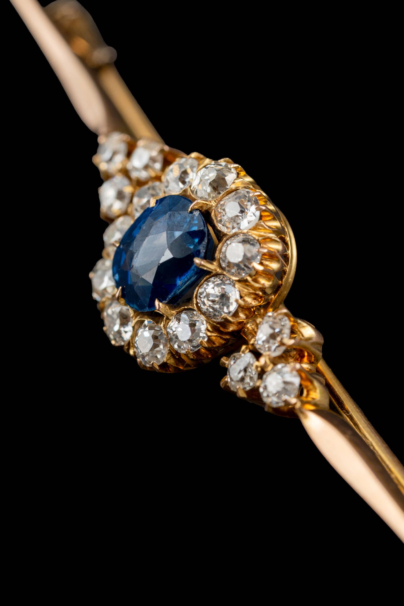 Dettaglio di una spilla vintage in oro con pietra blu centrale e pietre bianche incastonate, fotografia di gioielli su fondo nero